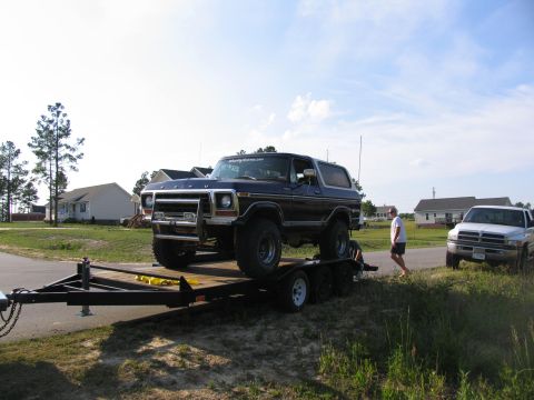 1995 Ford C-350 (Centurion Conversions) - A dark blue Ford Bronco is loaded onto a trailer, with a white pickup truck visible in the background.