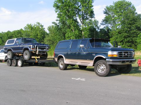 1995 Ford C-350 (Centurion Conversions) - A dark blue Ford Bronco is loaded onto a trailer, being towed by a dark green Ford F-Series SUV.