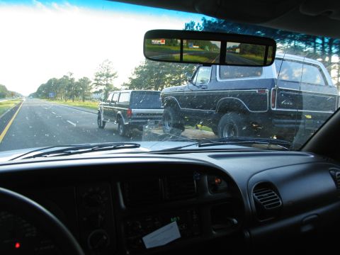 1995 Ford C-350 (Centurion Conversions) - A black Ford Bronco is towed on a trailer behind another vehicle on the highway.