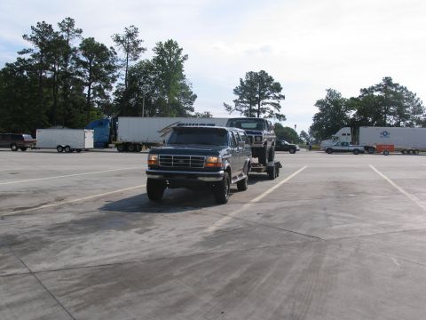 1995 Ford C-350 (Centurion Conversions) - A dark blue Ford F-Series truck tows a dark blue Ford Bronco on a trailer in a parking lot.