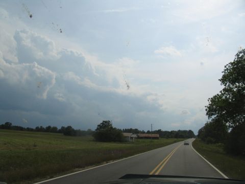 1995 Ford C-350 (Centurion Conversions) - A car travels down a rural road under a cloudy sky, with fields and trees lining the way.
