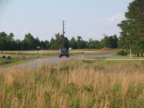 1978 Ford Bronco - A blue pickup truck approaches an intersection on a sunny day, with a red truck visible in the distance.