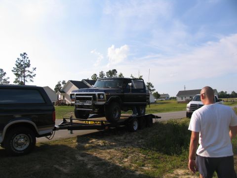 1978 Ford Bronco - A dark blue 1978 Ford Bronco is loaded onto a trailer, towed by another black Bronco, with houses in the background.