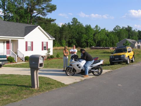 1978 Ford Bronco - People gather around a motorcycle in front of a house, with a yellow SUV visible on the street.