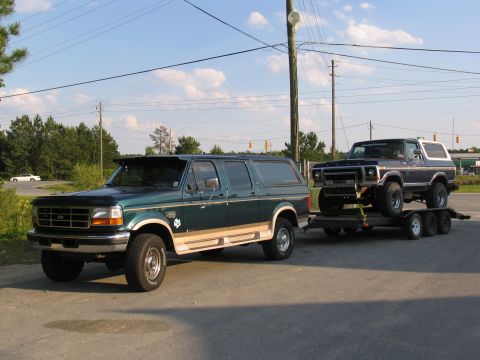 1978 Ford Bronco - A green Ford Bronco tows a blue Ford Bronco on a trailer under a bright sky.