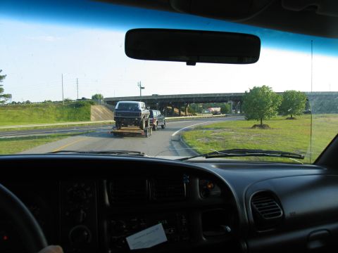 1978 Ford Bronco - A pickup truck tows a trailer with a dark SUV on the highway under an overpass.