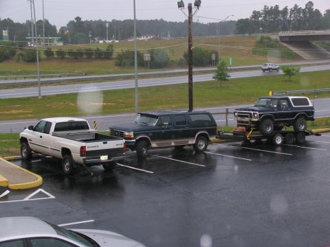 1978 Ford Bronco - A dark blue 1978 Ford Bronco is transported on a trailer, with a dark green SUV and a white Dodge pickup truck visible in the parking lot.