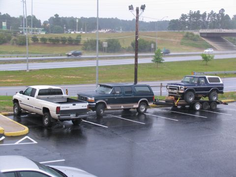 1978 Ford Bronco - A blue 1978 Ford Bronco is loaded onto a trailer, parked in a wet lot next to other vehicles.