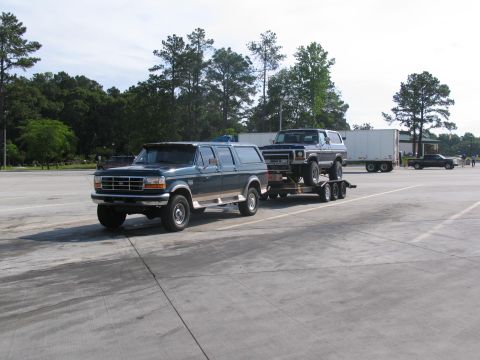 1978 Ford Bronco - A dark green SUV tows a blue SUV on a trailer in a parking lot.