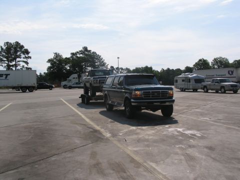 1978 Ford Bronco - A dark green SUV tows a pickup truck on a trailer in a parking lot under a partly cloudy sky.