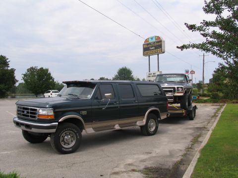 1978 Ford Bronco - A dark green SUV tows a dark blue pickup truck on a trailer near a steakhouse sign.