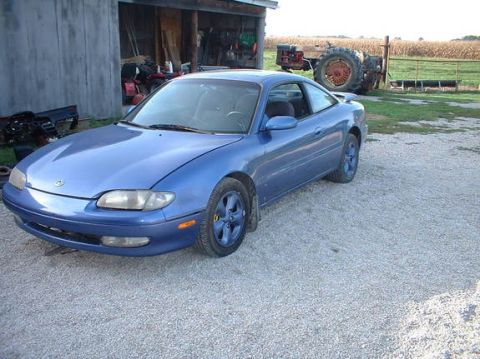 1995 Ford Probe - A blue coupe sits in a gravel lot next to a barn, with a tractor and cornfield visible in the distance.