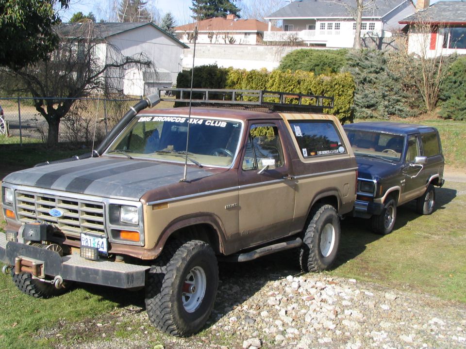 Brown lifted full-size Ford Bronco with roof rack and winch, next to a blue Bronco II on gravel.