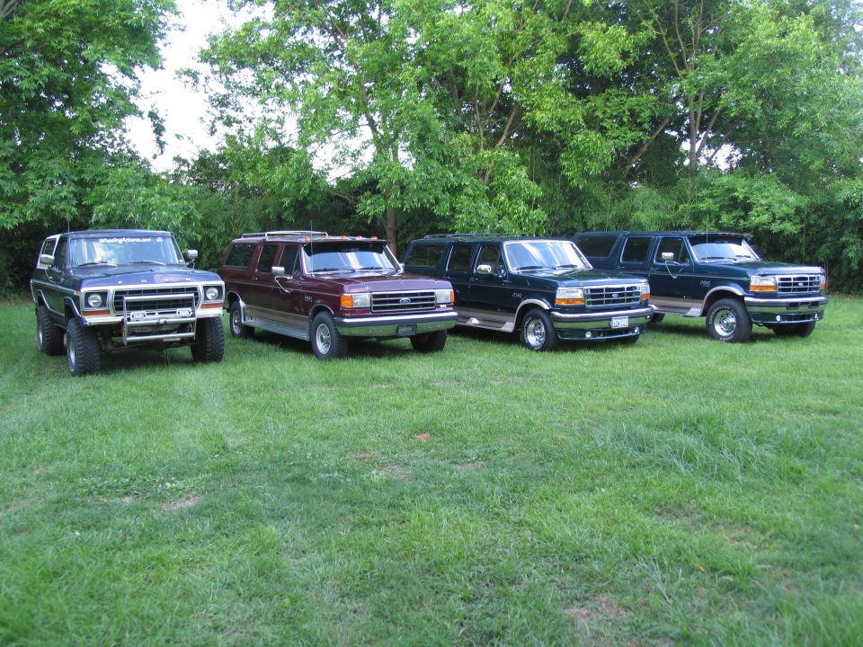 Four Ford SUVs parked on grass, three are dark green and one is maroon. The front of the leftmost SUV is visible