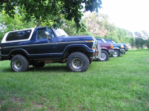 1995 Ford C-350 (Centurion Conversions) - A line of Ford Broncos and trucks are parked on a grassy field under trees.