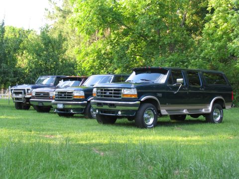 1995 Ford C-350 (Centurion Conversions) - Several Ford SUVs are lined up on a sunny day in a grassy field with trees behind them.