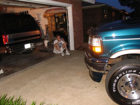 1995 Ford C-350 (Centurion Conversions) - Two Ford trucks, a teal F-Series and a black Bronco, are parked in a garage with a man sitting between them.