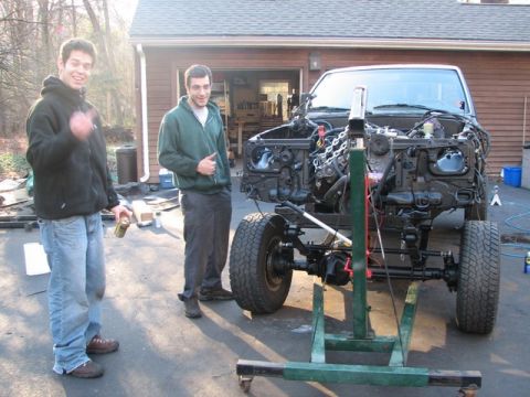 1994 Toyota Tacoma - A truck chassis is suspended by an engine hoist, with its engine removed. Two men are present in the driveway.