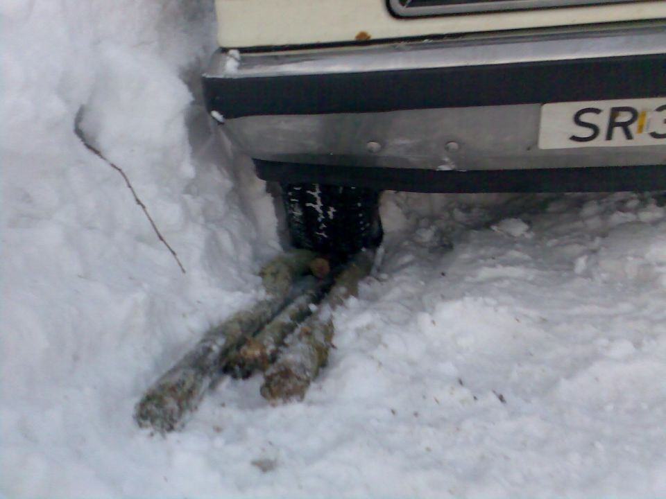 Rear bumper and tire of a vehicle stuck in snow, with logs under the tire