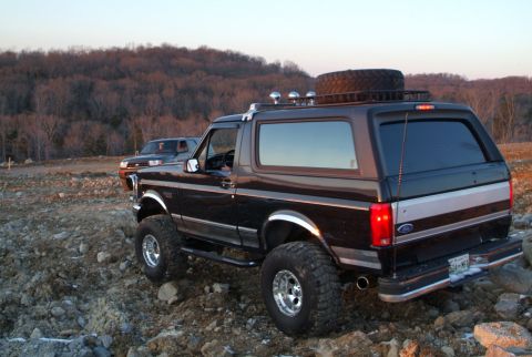 1995 Ford Bronco - Rear three-quarter view of a lifted black Ford Bronco parked on a rocky hillside at golden hour, showing the removable hardtop, chrome fend…