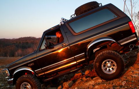 1995 Ford Bronco - Low-angle driver's-side shot of a black lifted Ford Bronco tilted nose-down on a rocky slope at sunset, with a driver visible behind the wh…