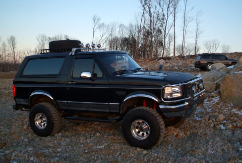 1995 Ford Bronco - A lifted black 1995 Ford Bronco sits on a rocky surface with trees in the background and another SUV parked on a hill.