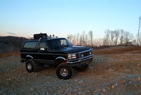 1995 Ford Bronco - This black 1995 Ford Bronco is equipped with a roof rack and spare tire, seen parked on a rocky terrain.