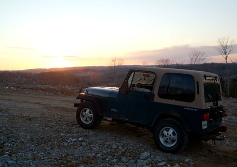1995 Ford Bronco - Dark blue Jeep Wrangler YJ with a tan soft top parked on a rocky graded hilltop at sunset, photographed from the driver's side in three-qua…