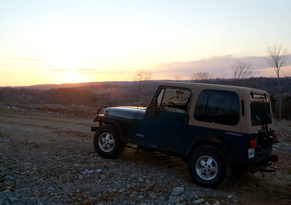 Blue Jeep Wrangler YJ with tan soft top on rocky hilltop at sunset