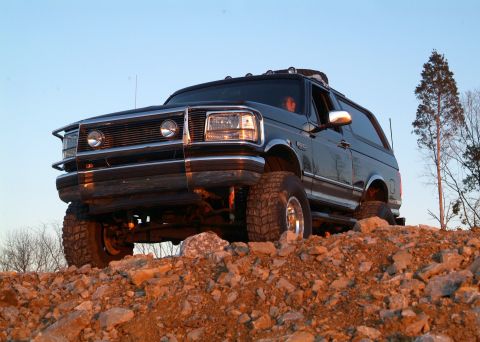 1995 Ford Bronco - Dramatic low-angle front three-quarter shot of a lifted black Ford Bronco perched on a rocky berm against a clear evening sky, with the sol…