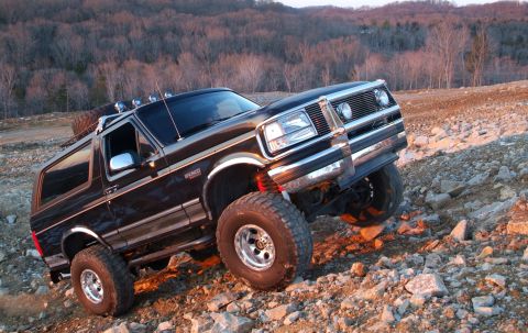 1995 Ford Bronco - Lifted black Ford Bronco flexing over a rock pile at an off-road site, front passenger tire climbing while the body leans, shot from a low…