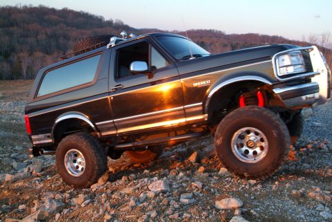 1995 Ford Bronco - Black lifted Ford Bronco flexing over a rocky hillside quarry at sunset, photographed from the driver's side in three-quarter front view wi…