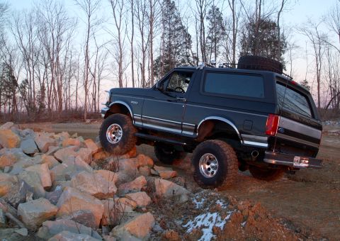 1995 Ford Bronco - Black lifted Ford Bronco climbing a pile of light-colored rocks at dusk, seen from the driver's side in three-quarter rear view with the fr…