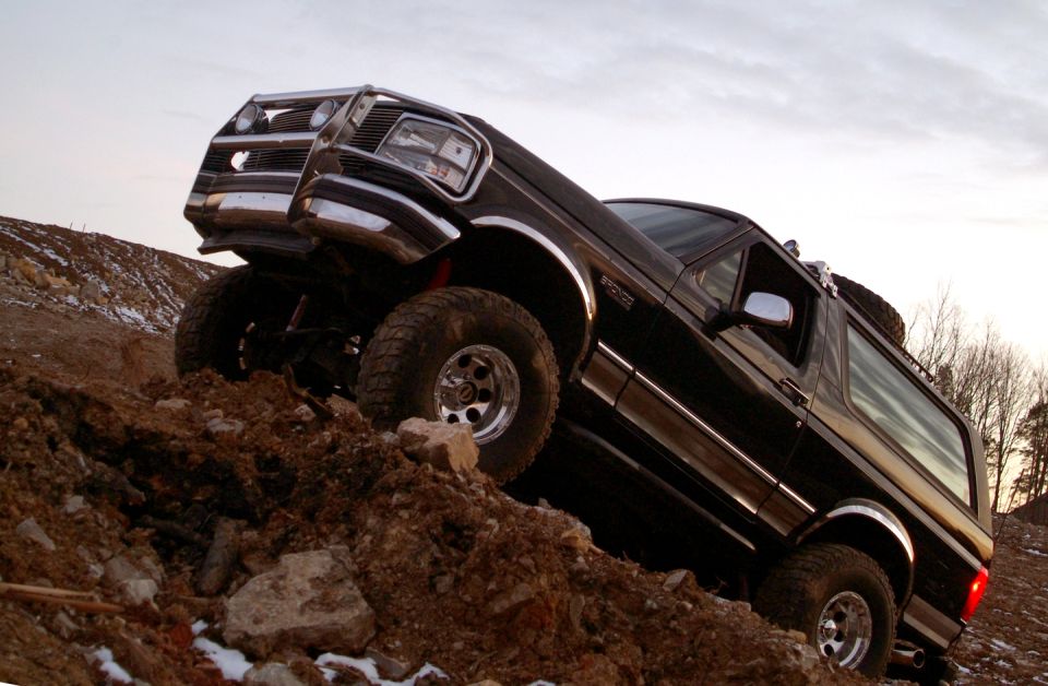 Black Ford Bronco climbing a dirt hill, front-left quarter view, modified with large tires and aftermarket grille guard