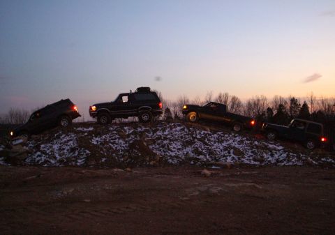 1995 Ford Bronco - Four 4x4s posed at dusk across a snow-patched dirt mound, silhouetted against an orange-and-blue sunset sky: an SUV nosing down the left sl…