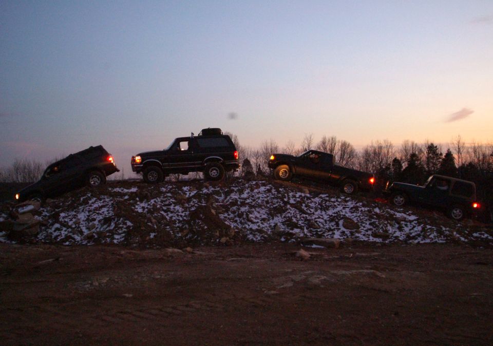 Four 4x4s including a lifted Ford Bronco posed on a snowy dirt mound at sunset