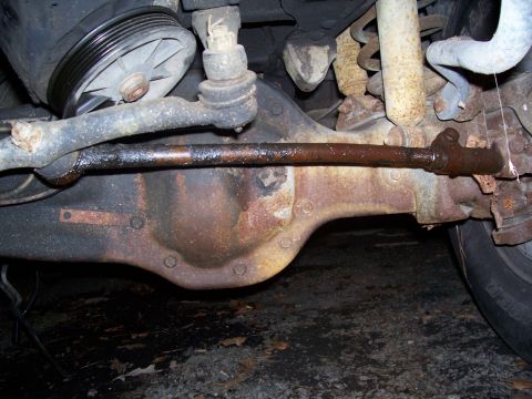 1985 Ford Bronco - Close-up view of the front axle and steering components, showing a rusty tie rod and suspension parts.