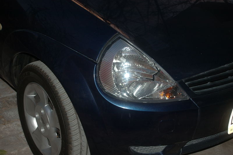 Night close-up of dark blue hatchback front corner, clear headlamp and silver five-spoke wheel