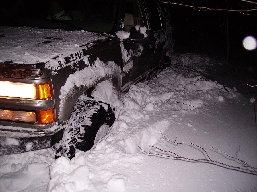 Front corner of a dark SUV, covered in snow, with its front tire deep in a snowdrift. Headlight is on