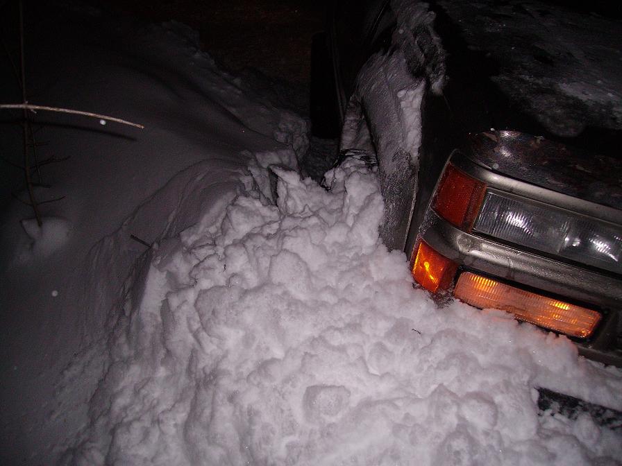 Vehicle front corner stuck deep in snow, headlights and turn signals visible