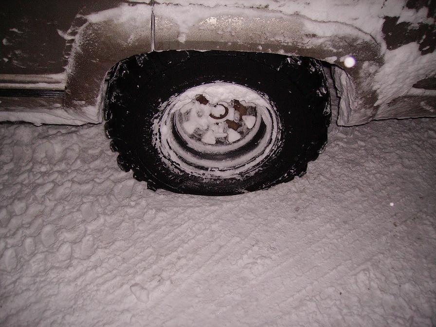Close up of a vehicle's tire covered in snow, with snow on the vehicle's body and tire tracks in the snow on the ground