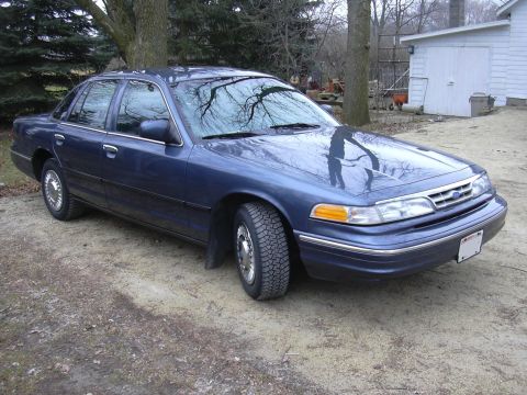 1996 Ford Crown Victoria - A blue sedan is parked on a dirt lot, with trees and a white building visible.