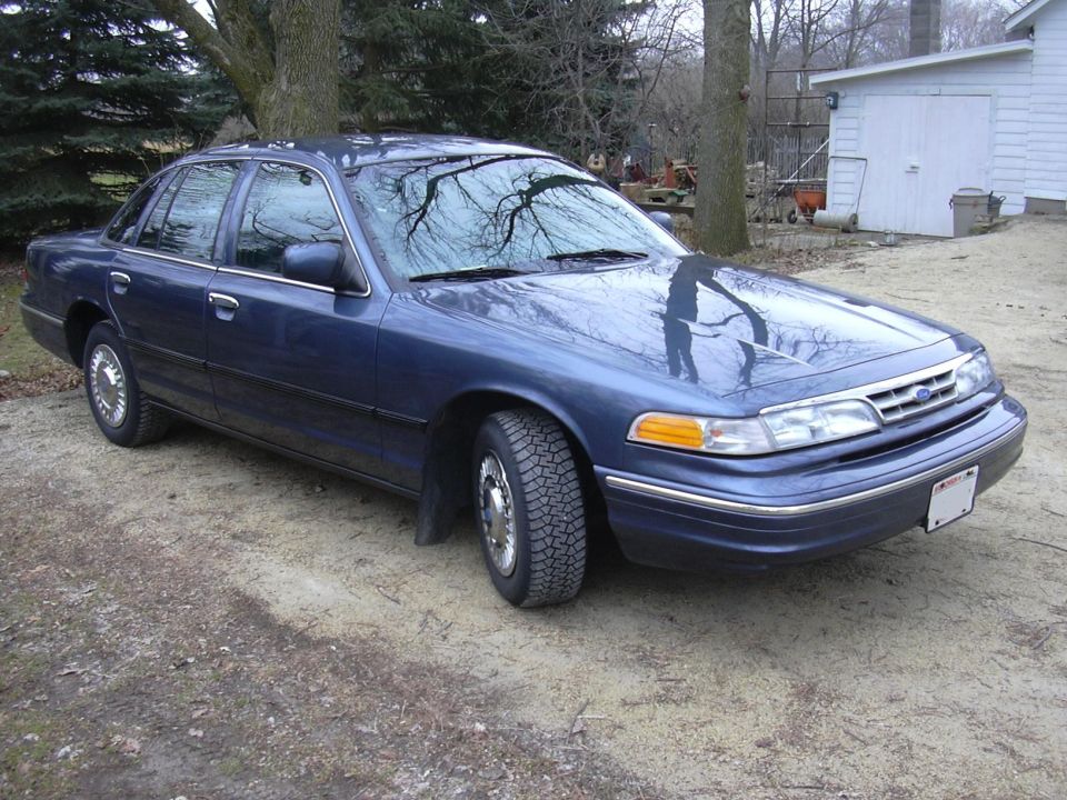Blue sedan parked on dirt lot with trees and a white building in the background