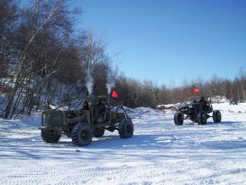 2002 Jeep Wrangler - Two off-road vehicles drive through a snow-covered area with trees in the background.