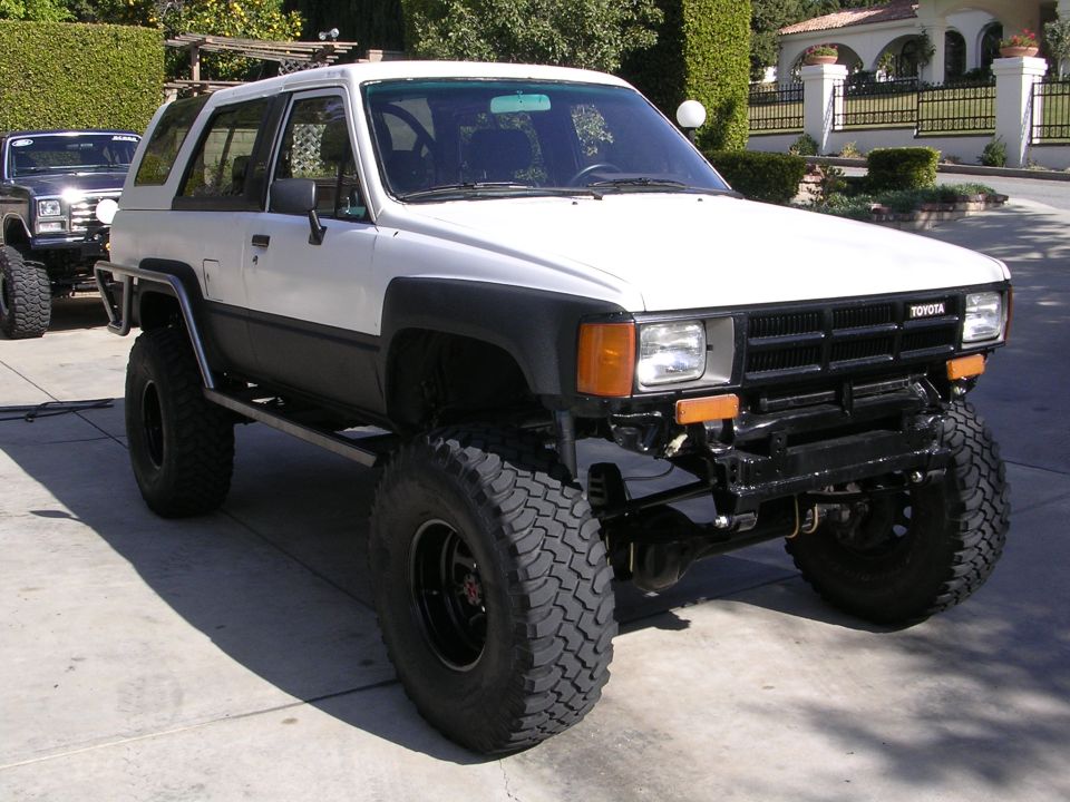 White Toyota 4Runner with big tires and custom bumper. Another dark SUV is parked behind it