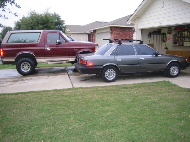 Lifted burgundy Ford Bronco parked in a driveway next to a gray Toyota sedan with a roof rack