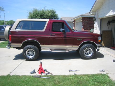 1996 Ford Bronco - A maroon 1996 Ford Bronco with a tan removable top is parked in a driveway with a jack and car parts nearby.