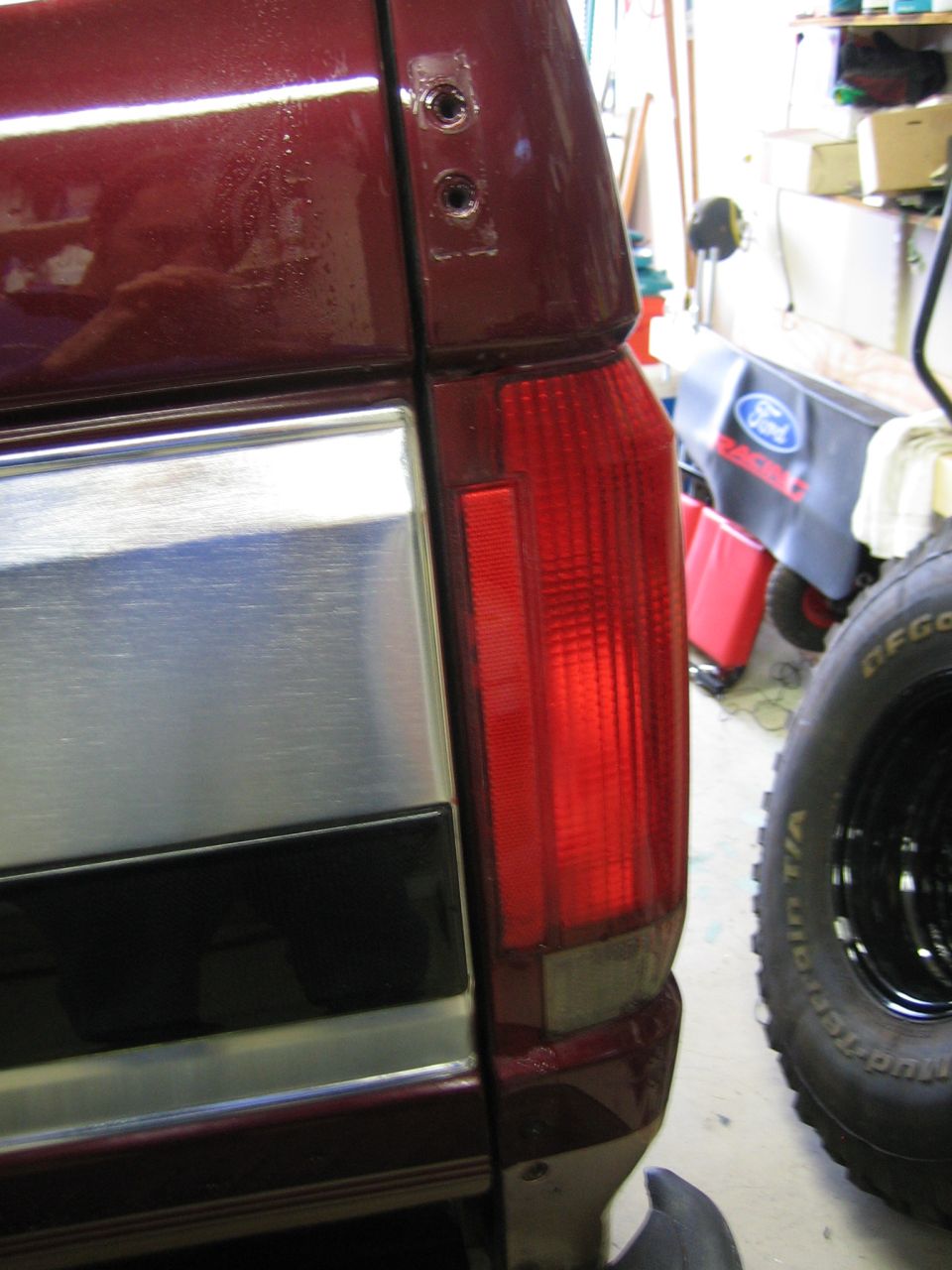 Close-up of a dark red SUV's rear corner, showing the taillight and a chrome strip. A tire is visible to the right