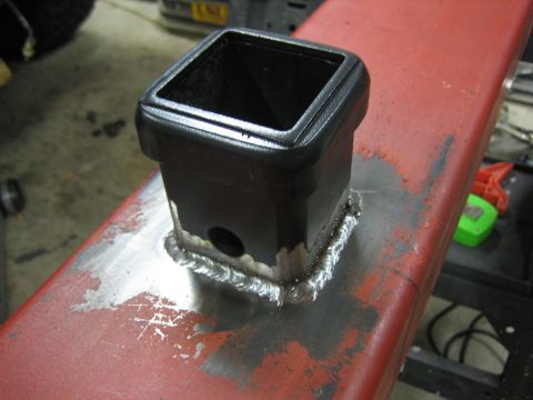 1996 Ford Bronco - A trailer hitch receiver is welded onto a red metal beam in a workshop setting.