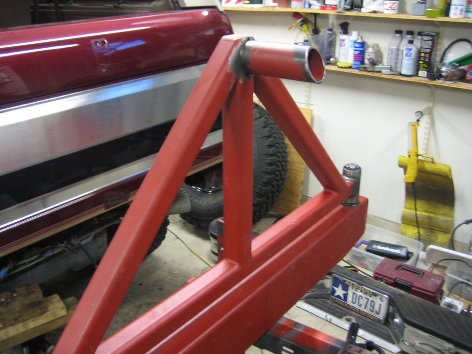 Red metal swing-away tire carrier frame attached to a red vehicle. A spare tire is visible behind the carrier